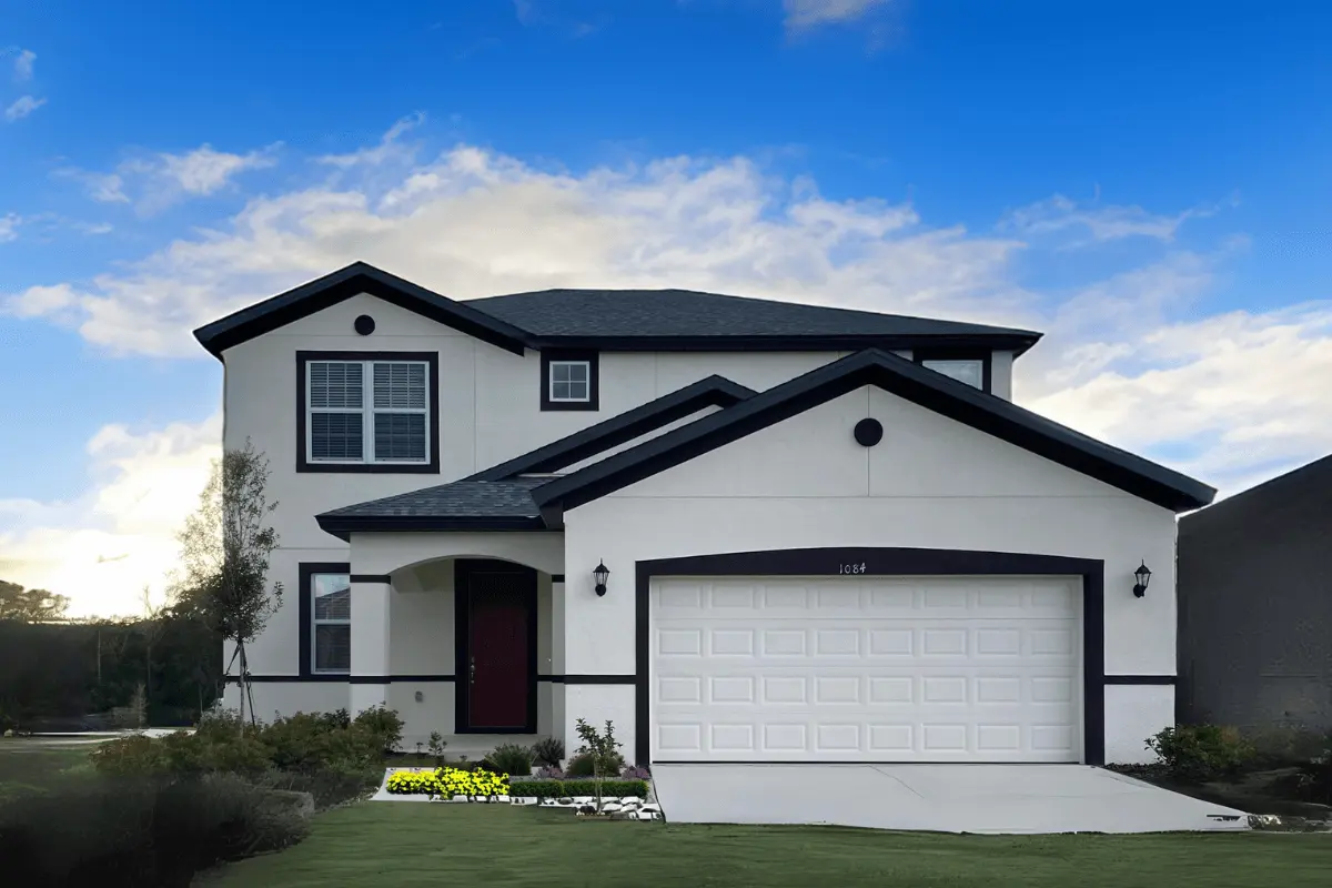 A two-story modern house with white walls, black trim, a red door, and a double garage. There are flower beds, bushes, and a small tree in the front yard under a partly cloudy blue sky.