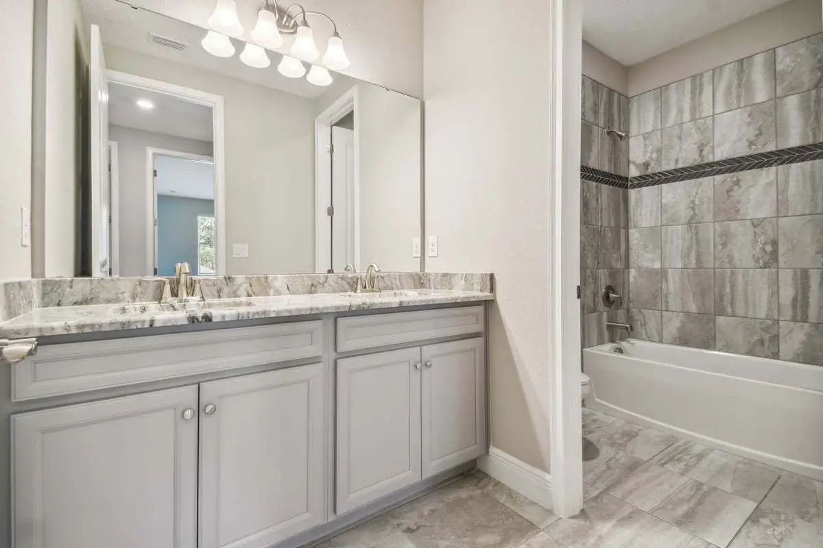 Modern bathroom with a large mirror above a double-sink vanity, light gray cabinets, marble countertop, and a bathtub with gray tile surround. The floor is tiled, and the walls are painted a light color.