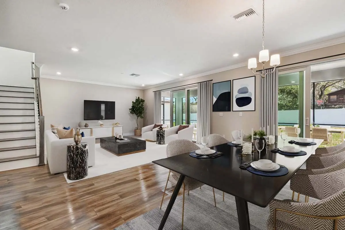 Modern open-plan living and dining area with light walls, wood floors, neutral furniture, wall art, large TV, dining table set for six, and glass doors opening to a green outdoor space.