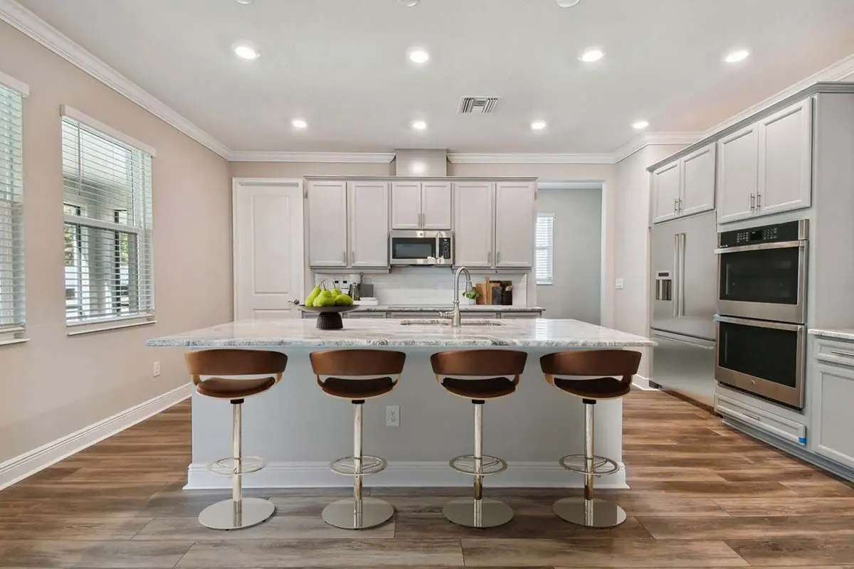 Modern kitchen with light gray cabinets, marble island countertop, four brown barstools, stainless steel appliances, wooden flooring, and recessed ceiling lights. Three windows let in natural light on the left side.