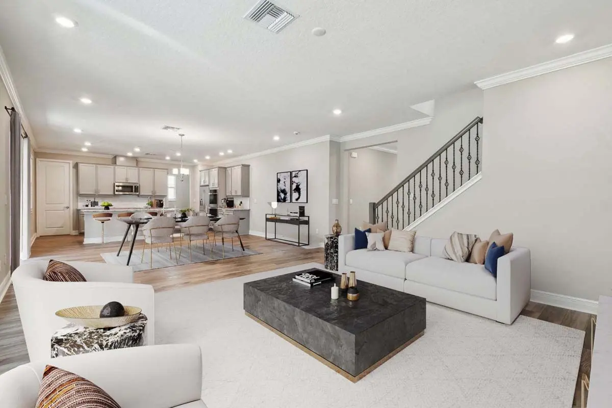 Modern open-concept living room and kitchen with neutral tones, white couches, a dark coffee table, dining area with gray chairs, and a staircase with a decorative railing. Bright, well-lit space with wood and tile flooring.