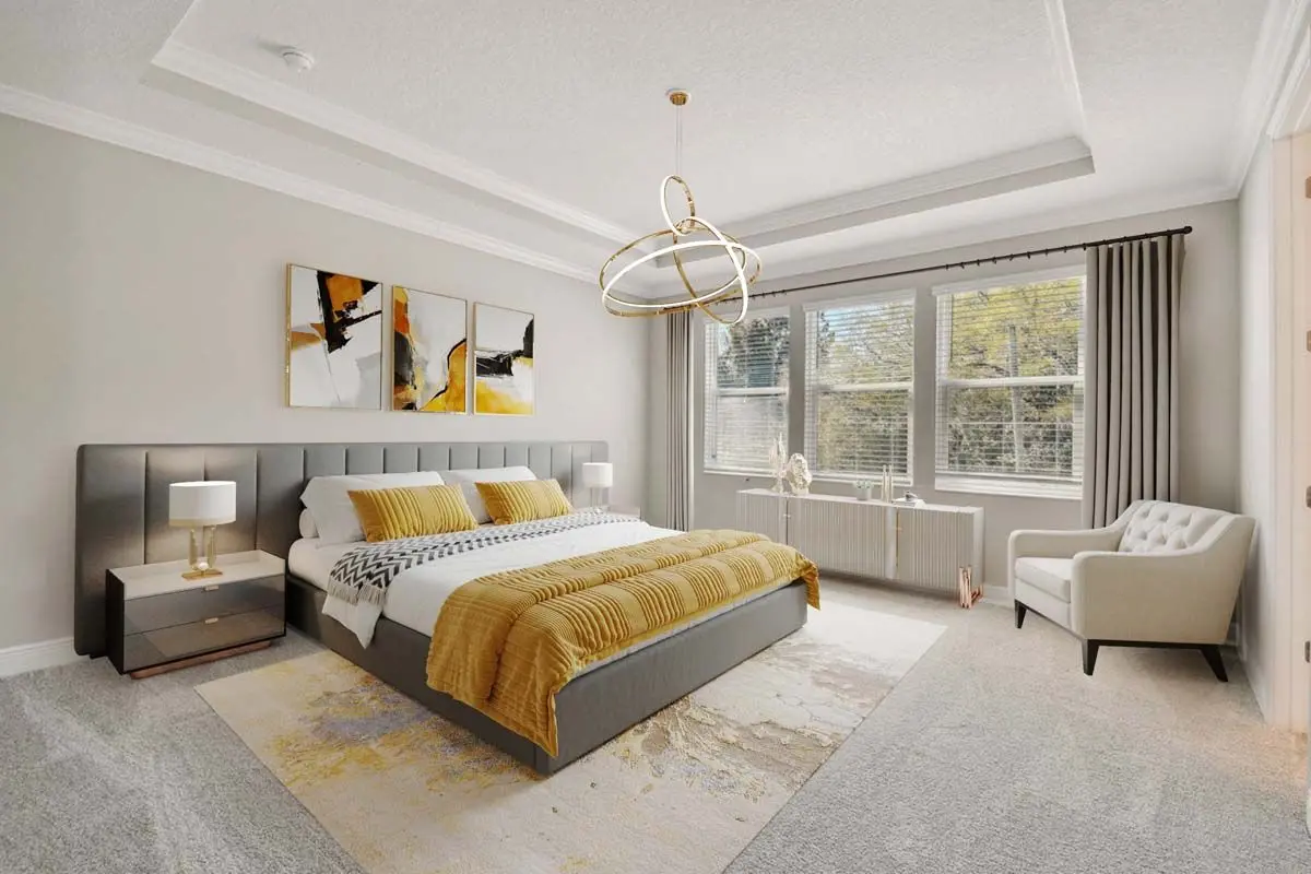 Modern bedroom with gray walls, a large bed with yellow accents, abstract artwork above the headboard, two nightstands, a gold chandelier, a white armchair by the window, and natural light streaming in.