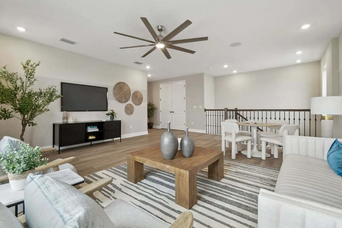 Modern living room with light walls, wood flooring, and neutral decor. Features a large ceiling fan, a striped rug, a wooden coffee table with vases, a sofa, armchairs, and a TV on a console. Dining area in the background.