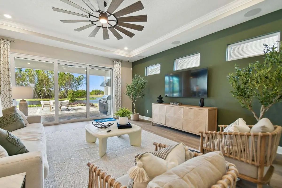 Modern living room with green accent wall, wooden furniture, large flat-screen TV, indoor plants, beige sofa and chairs, white coffee table, ceiling fan, and sliding glass doors opening to a patio with outdoor seating.