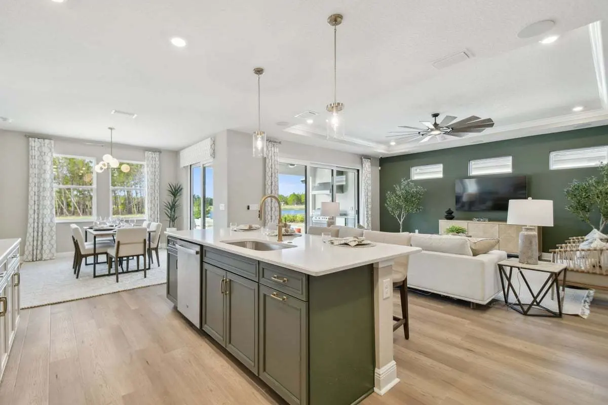 Modern open-concept kitchen and living area with an island, pendant lights, green accent cabinets, a white sectional sofa, large windows, and a dining table near sliding glass doors with outdoor views.