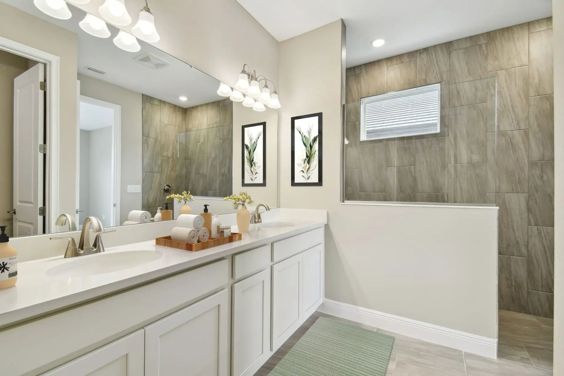 Modern bathroom with a double sink vanity, large wall mirror, light fixtures above, two framed botanical prints, gray tiled shower with a window, and a green floor mat. Towels and soap are neatly arranged on the counter.