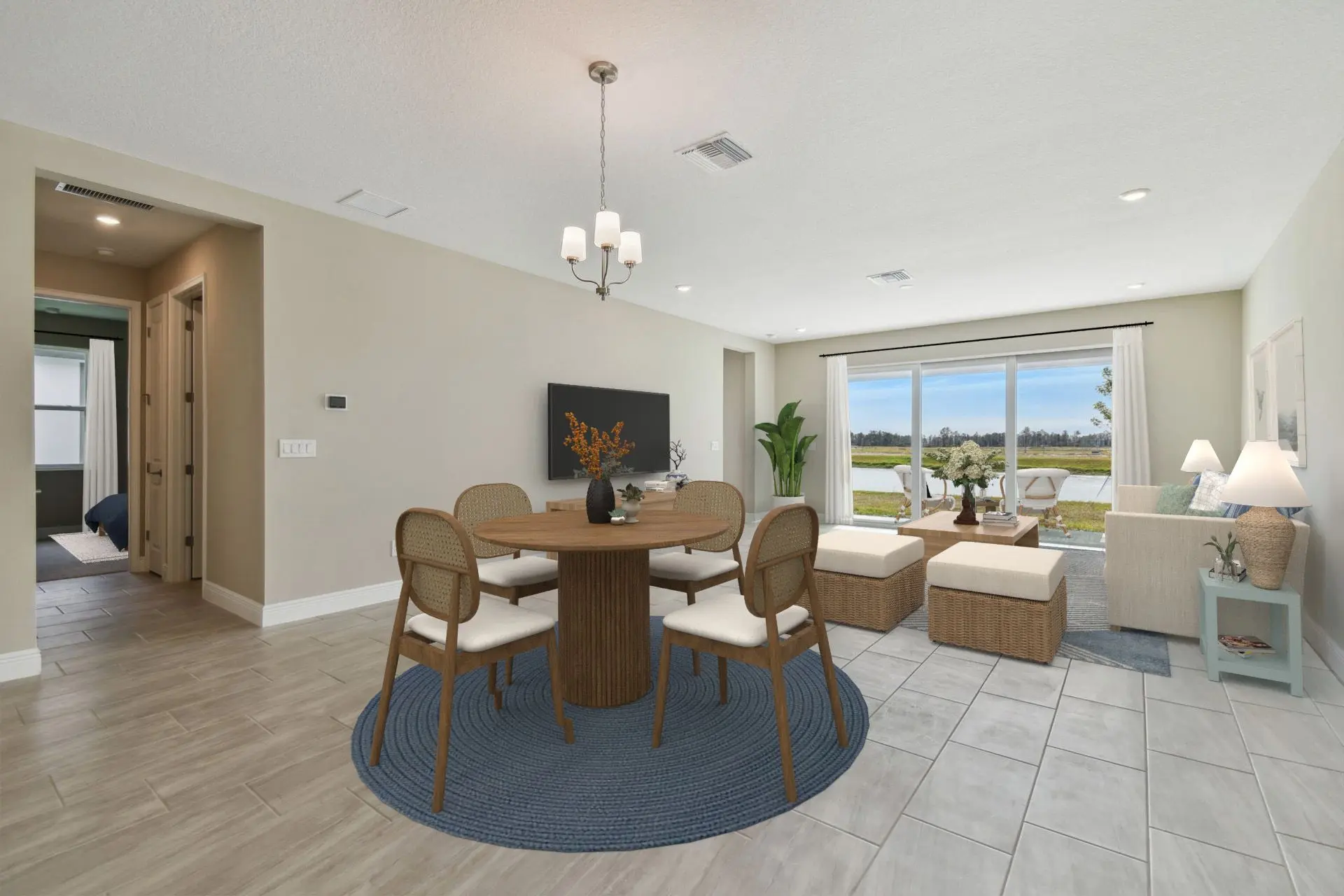 Modern open-concept living and dining room with beige walls, tile floors, round dining table with chairs, wicker furniture in the living area, large window with scenic outdoor view, and neutral decor.