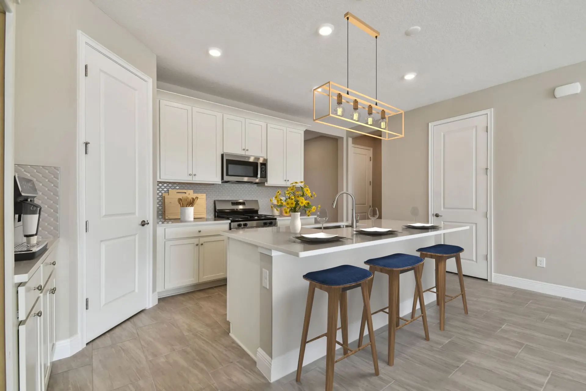 Modern kitchen with white cabinets, stainless steel appliances, a center island with three blue barstools, and a gold geometric pendant light. A vase of yellow flowers sits on the island.