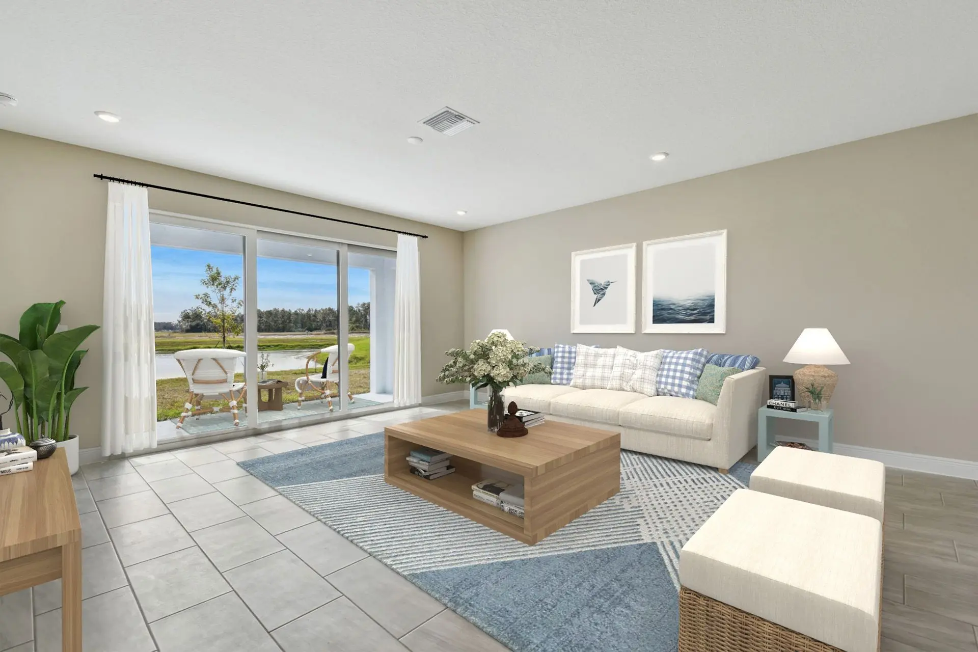 Bright living room with a large glass sliding door opening to a patio with chairs, a beige sofa, coffee table, blue rug, indoor plant, and light neutral decor. Outdoor view of greenery and water.