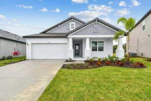 A modern, single-story suburban house with a gray exterior, a double garage, a front porch with columns, manicured lawn, and landscaping, under a blue sky with scattered clouds.