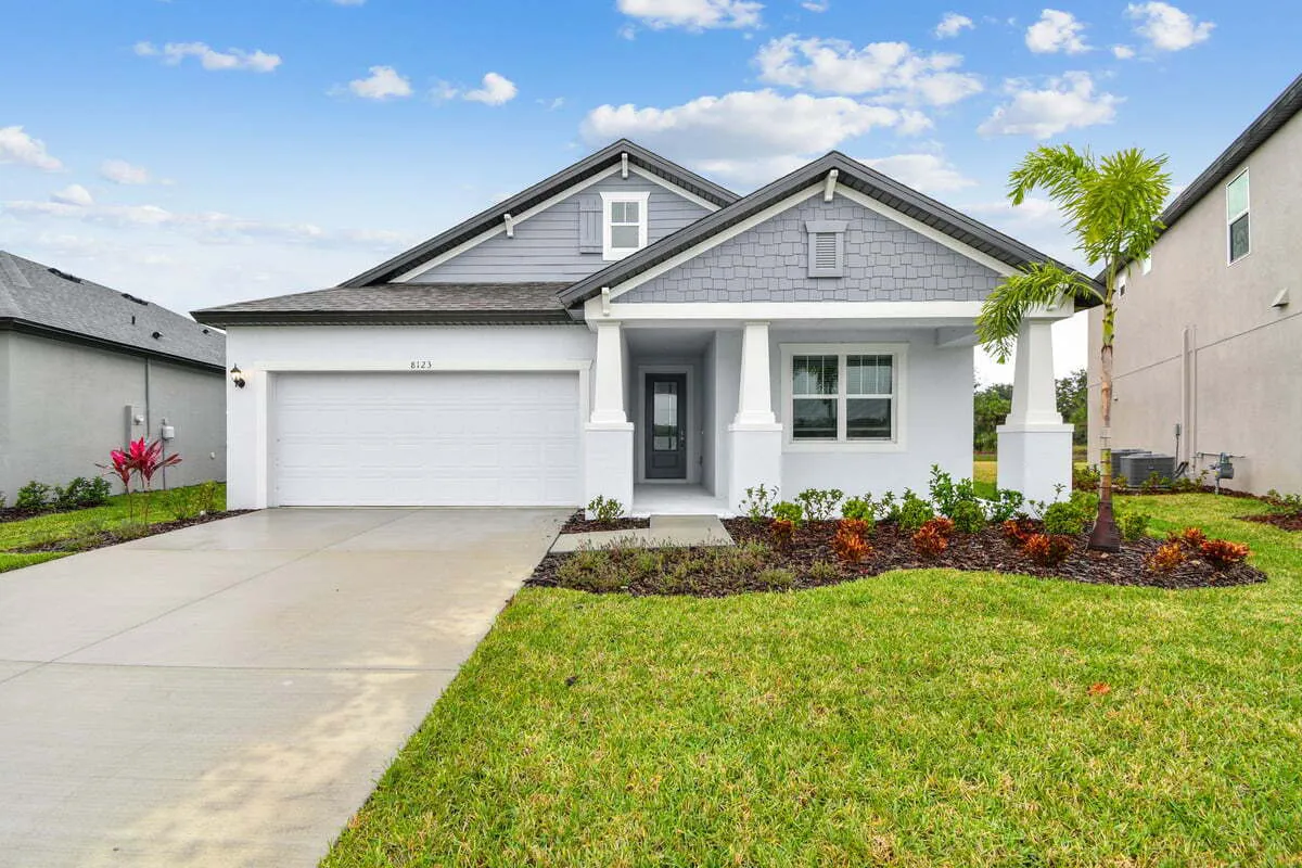 A modern, single-story suburban house with a gray exterior, a double garage, a front porch with columns, manicured lawn, and landscaping, under a blue sky with scattered clouds.