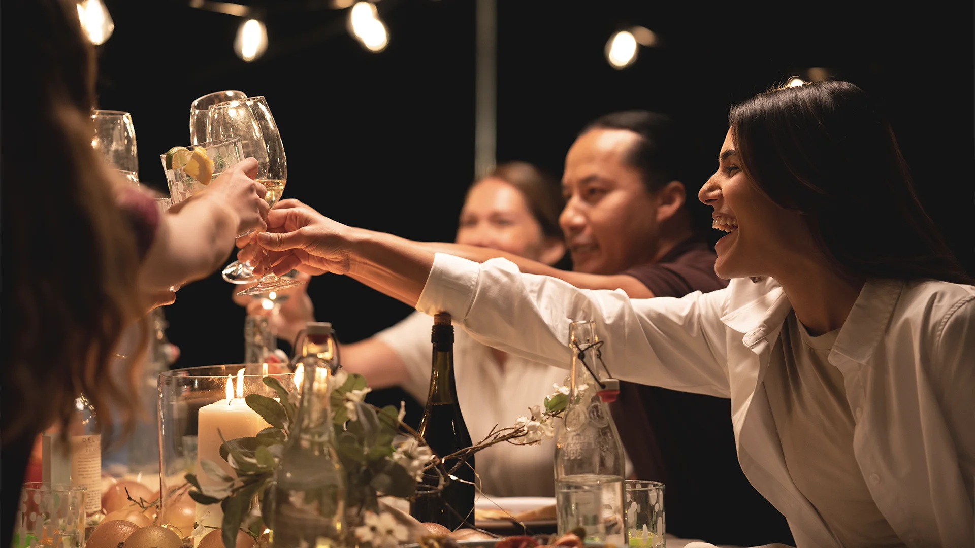 A group of people sit around a table outdoors at night, smiling and toasting with glasses. The table is decorated with candles, flowers, and bottles. String lights glow softly in the background.