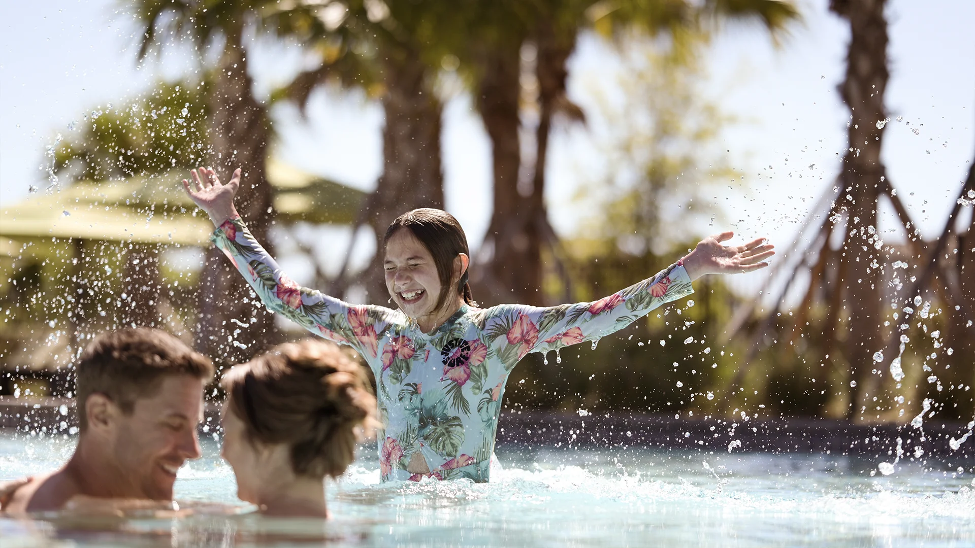Young girl in a floral swimsuit splashes water with arms raised and smiles in a pool, while two adults face each other in the foreground. Palm trees and bright sunlight create a vibrant, joyful atmosphere.