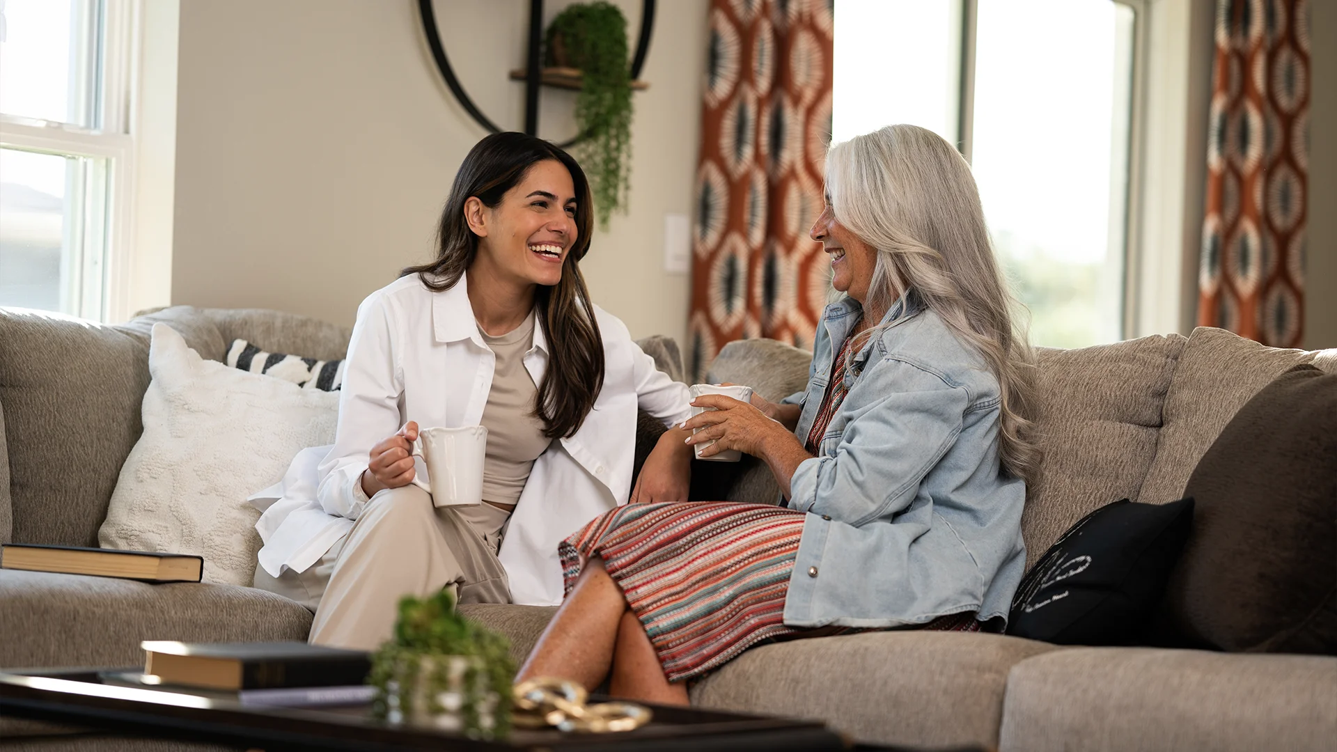 A younger woman in a white coat and an older woman with long gray hair sit on a couch, smiling and talking while holding mugs in a cozy, well-lit living room.