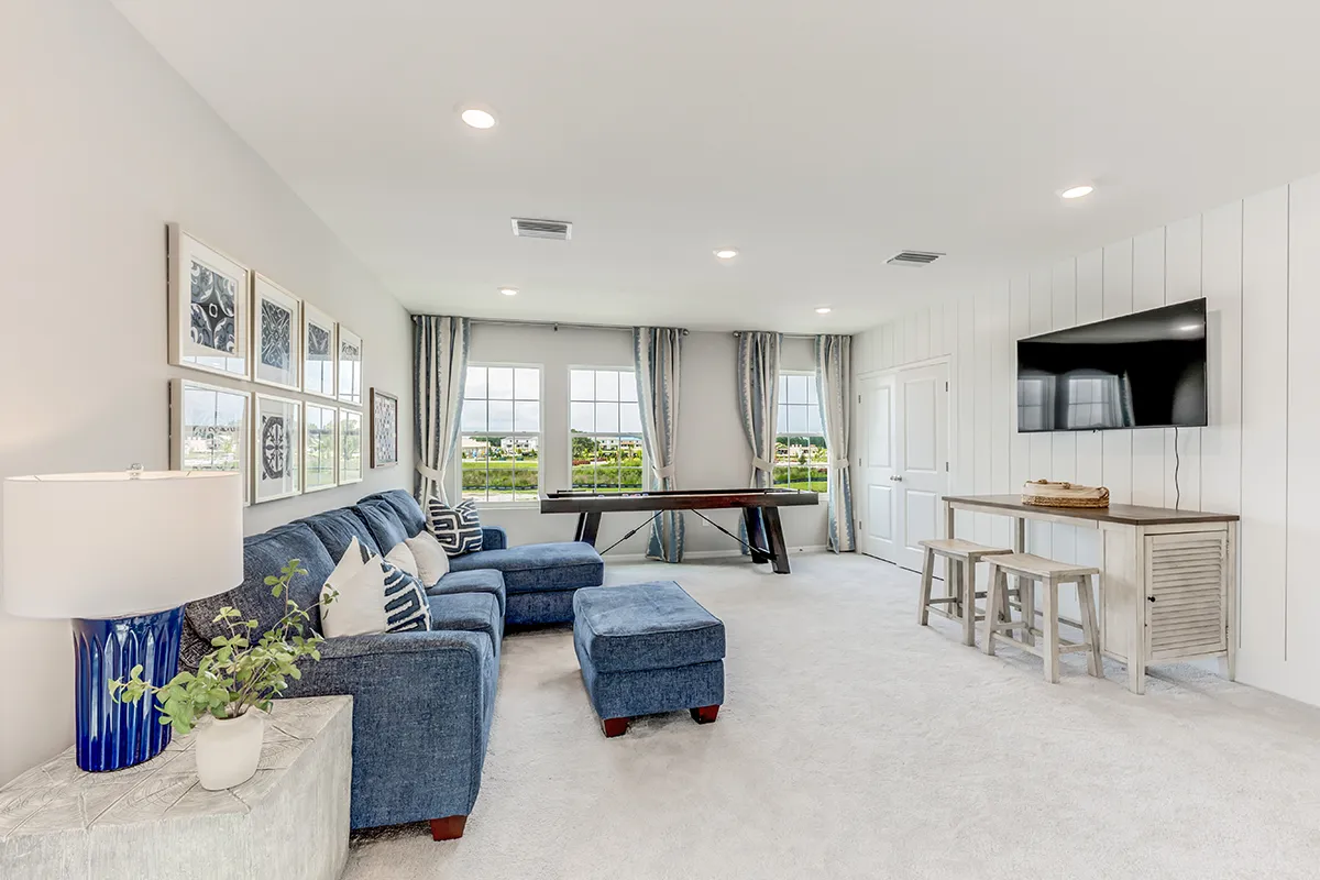 Bright living room with blue sectional sofa, wall art, shuffleboard table by large windows, flat-screen TV mounted above a console table, and neutral decor on a light carpeted floor.