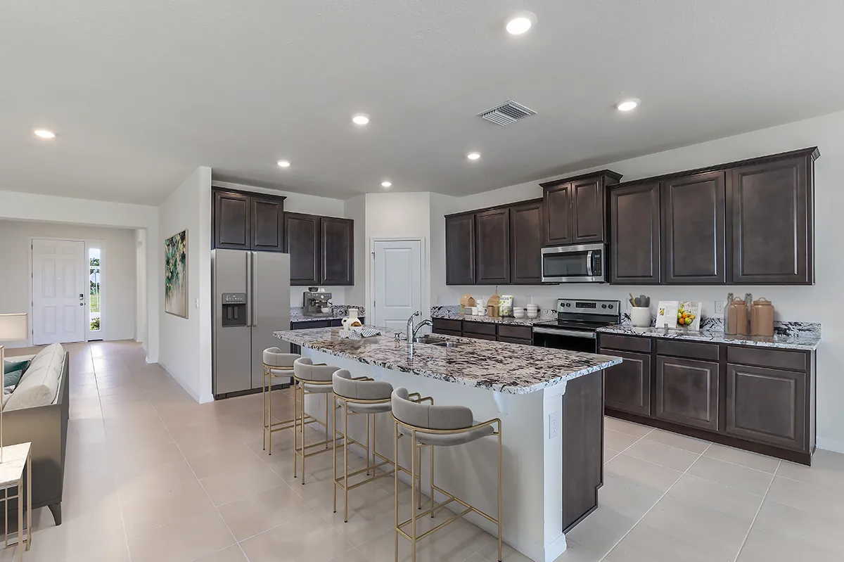 Modern kitchen with dark wood cabinets, stainless steel appliances, and a large granite island with four barstools. The space is well-lit with recessed lighting and has tile flooring. A living area is visible to the left.