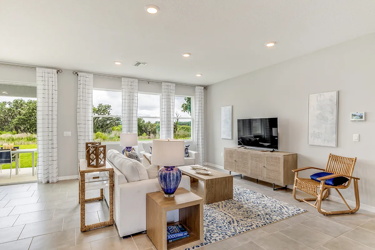 Bright living room with large windows, white sofa, blue accents, a rug, wooden coffee and side tables, TV on a console, wicker chair, tiled floor, and view of a green outdoor area.