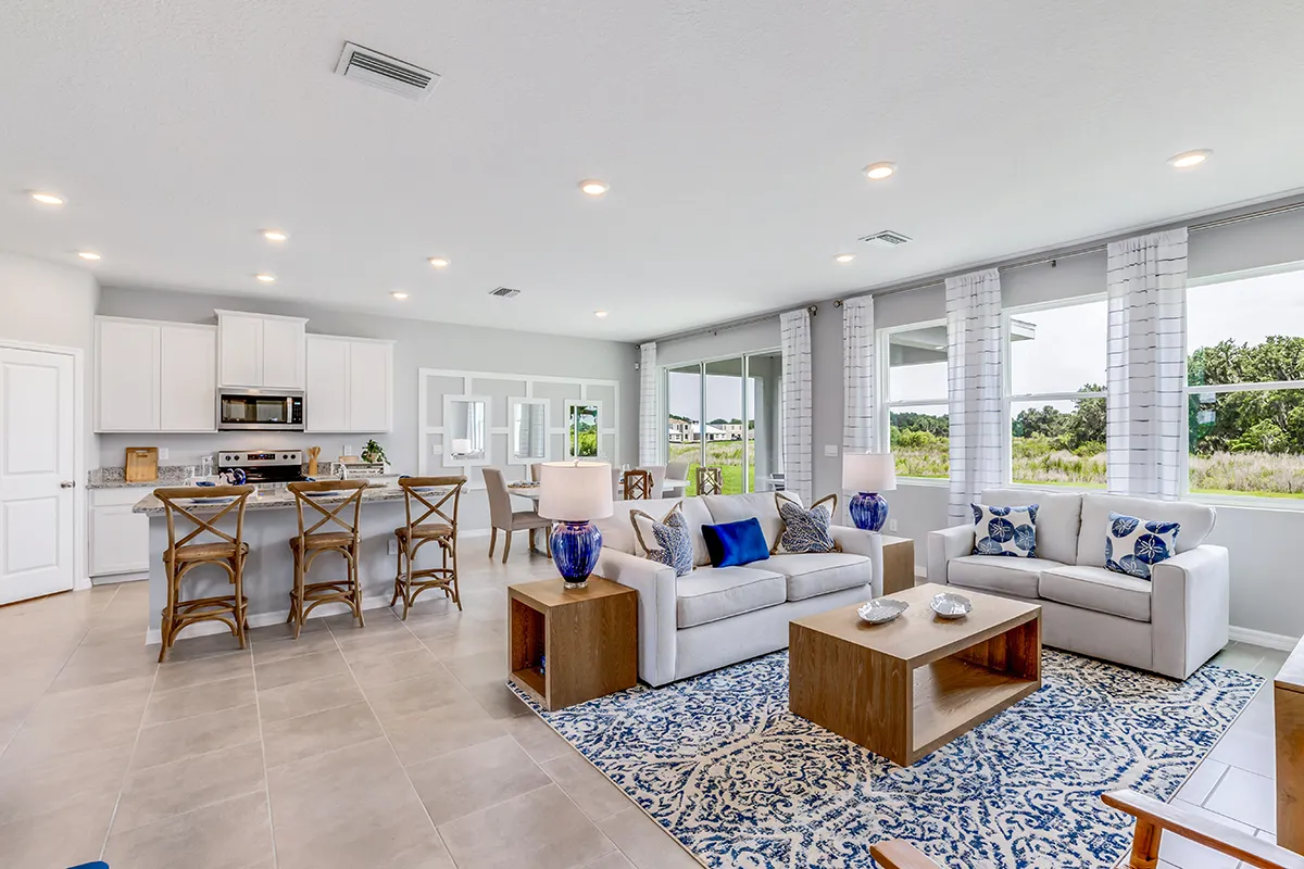 Open-concept living area with a kitchen, dining, and living room. Modern decor features blue and white accents, wooden furniture, bar stools, large windows with sheer curtains, and a view of greenery outside.