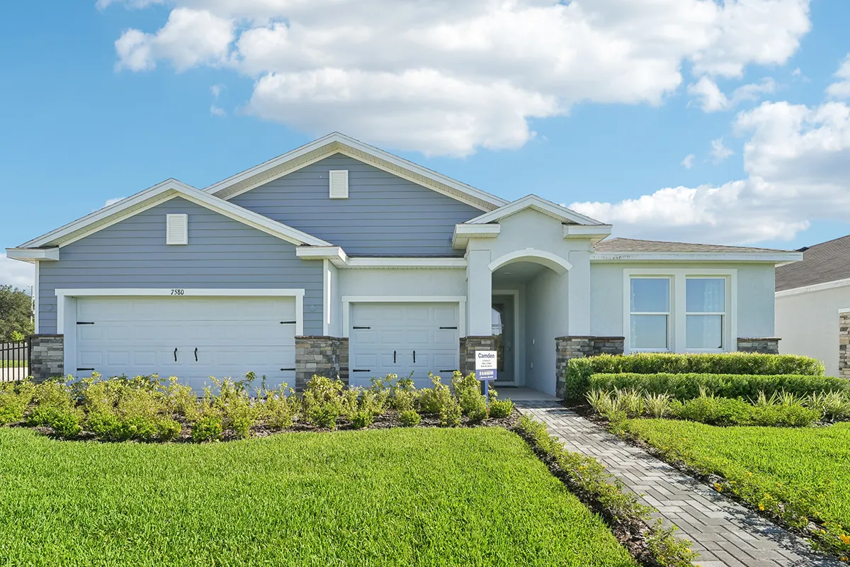 A modern single-story house with blue-gray siding, white trim, and three garage doors, surrounded by a green lawn and landscaped bushes under a partly cloudy sky.
