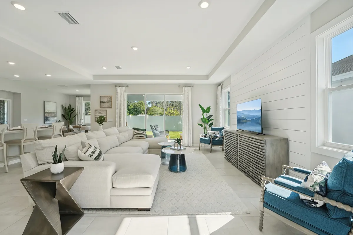 Bright, modern living room with a large white sectional sofa, coffee table, TV on a console, blue accent chairs, potted plants, and large windows letting in natural light. Open dining area is visible in the background.