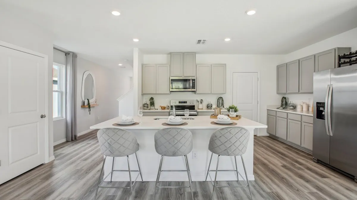 Modern kitchen with light gray cabinets, a stainless steel refrigerator, oven, and microwave. A white island with three cushioned stools is set with plates and napkins. The floor is light wood, and lighting is bright.