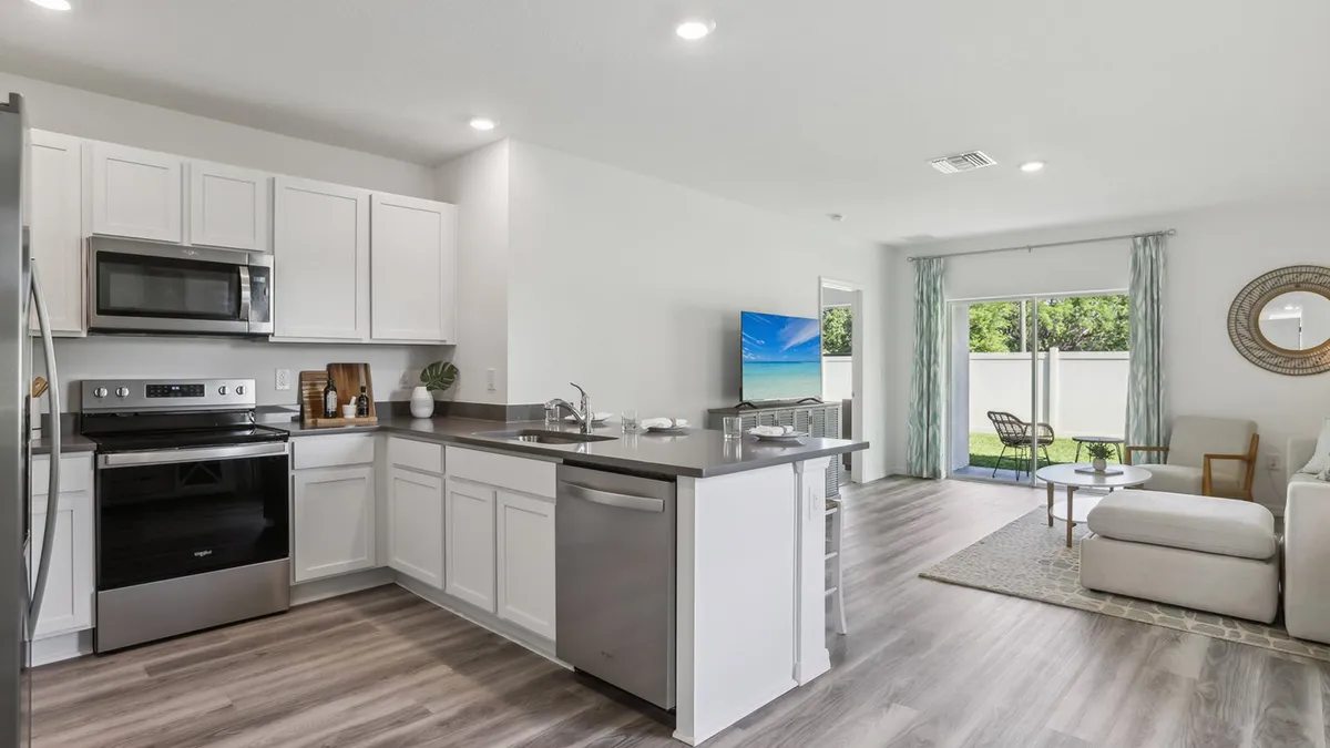 Modern open-concept kitchen and living room with white cabinets, stainless steel appliances, gray countertops, a sofa, TV, round mirror, and large sliding glass doors leading to a patio with greenery outside.