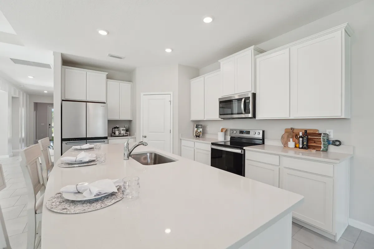 Modern kitchen with white cabinets, stainless steel appliances, and a large island with place settings for three. The space is bright with recessed lighting and a clean, minimalist design.