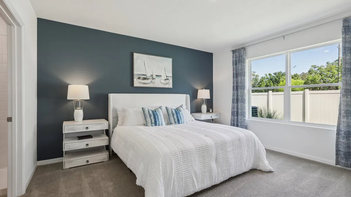 A bright bedroom with a blue accent wall, a bed with white bedding and blue pillows, two bedside tables with lamps, a sailboat painting above the bed, and a large window with blue curtains.