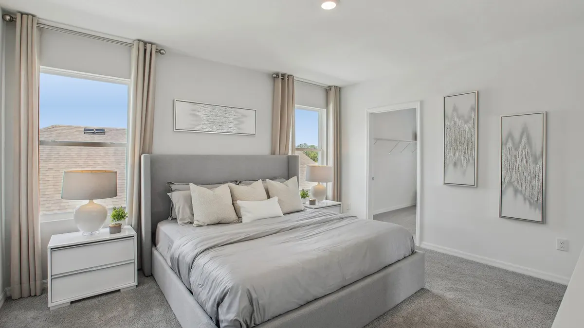 Modern bedroom with a gray upholstered bed, neutral bedding, two nightstands with lamps and plants, large windows with light curtains, minimal wall art, and an open walk-in closet.