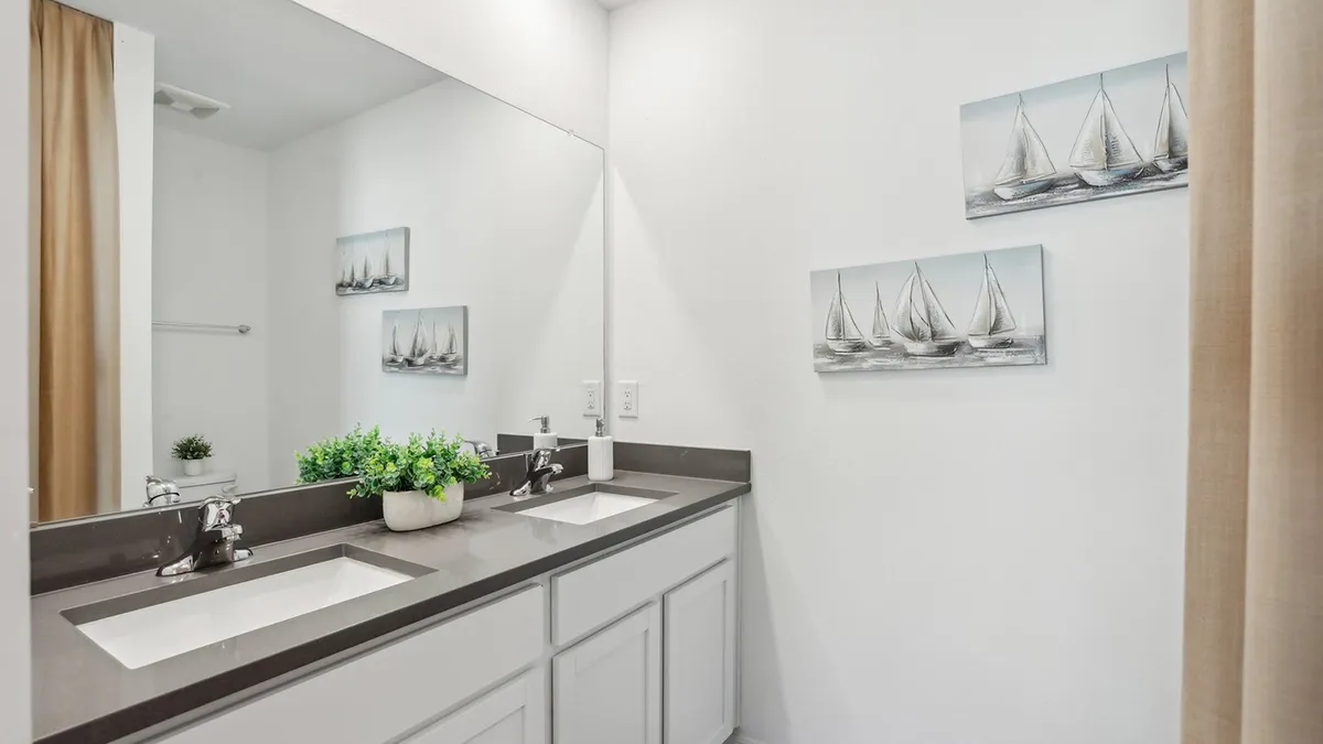 Modern bathroom with a double sink vanity, gray countertop, large mirror, potted plant, and two wall art pieces depicting sailboats on a white wall. Beige shower curtain partially visible on the right.