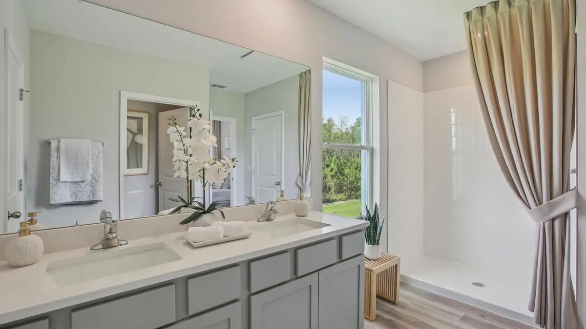 Modern bathroom with a double sink vanity, large mirror, white countertops, an orchid plant, a walk-in shower with beige curtains, a window, and a wooden stool holding a potted plant.