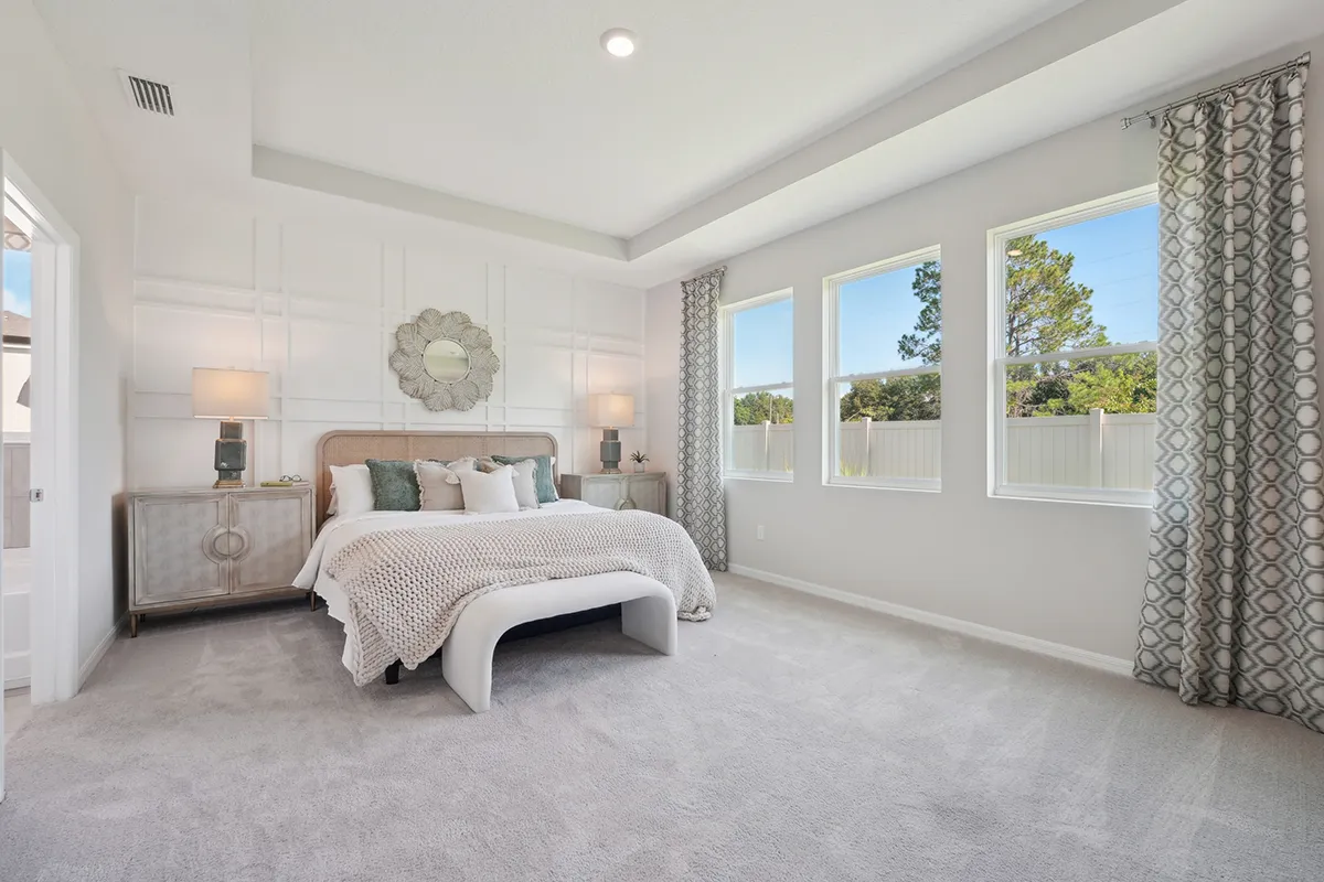 A bright, spacious bedroom with a large bed, two nightstands with lamps, patterned curtains, three tall windows letting in natural light, and light-colored carpeting. The decor is modern and neutral-toned.