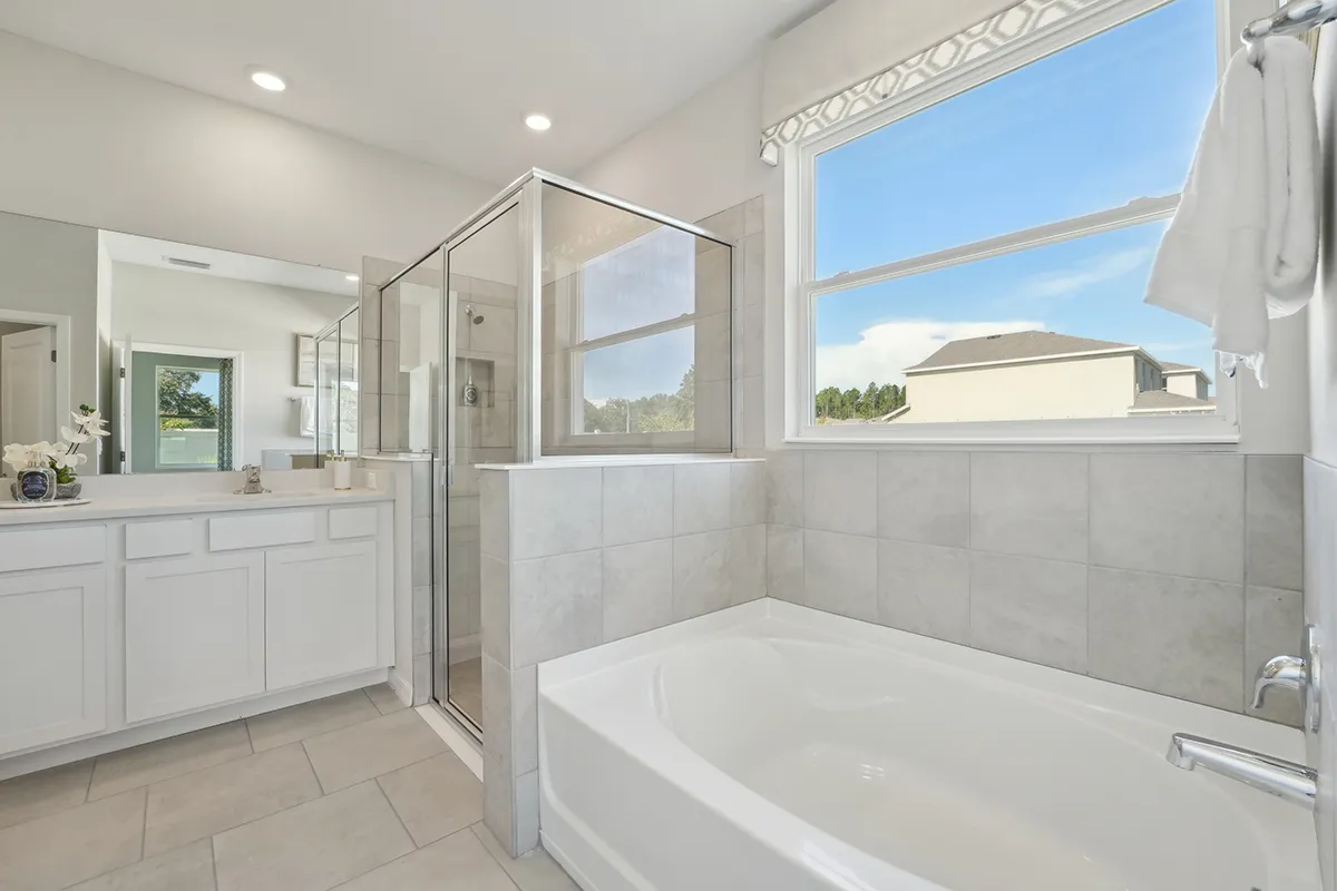Modern bathroom with a glass-enclosed shower, white bathtub, large window with a view outside, and a white vanity with a wide mirror, all in a neutral color scheme.