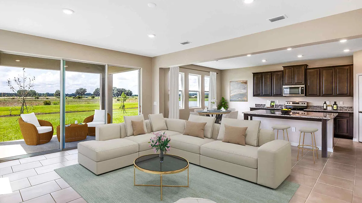 A modern, open-concept living room and kitchen with a cream sectional sofa, round coffee table, and large windows overlooking a green field. The kitchen features dark cabinets and a breakfast bar with stools.