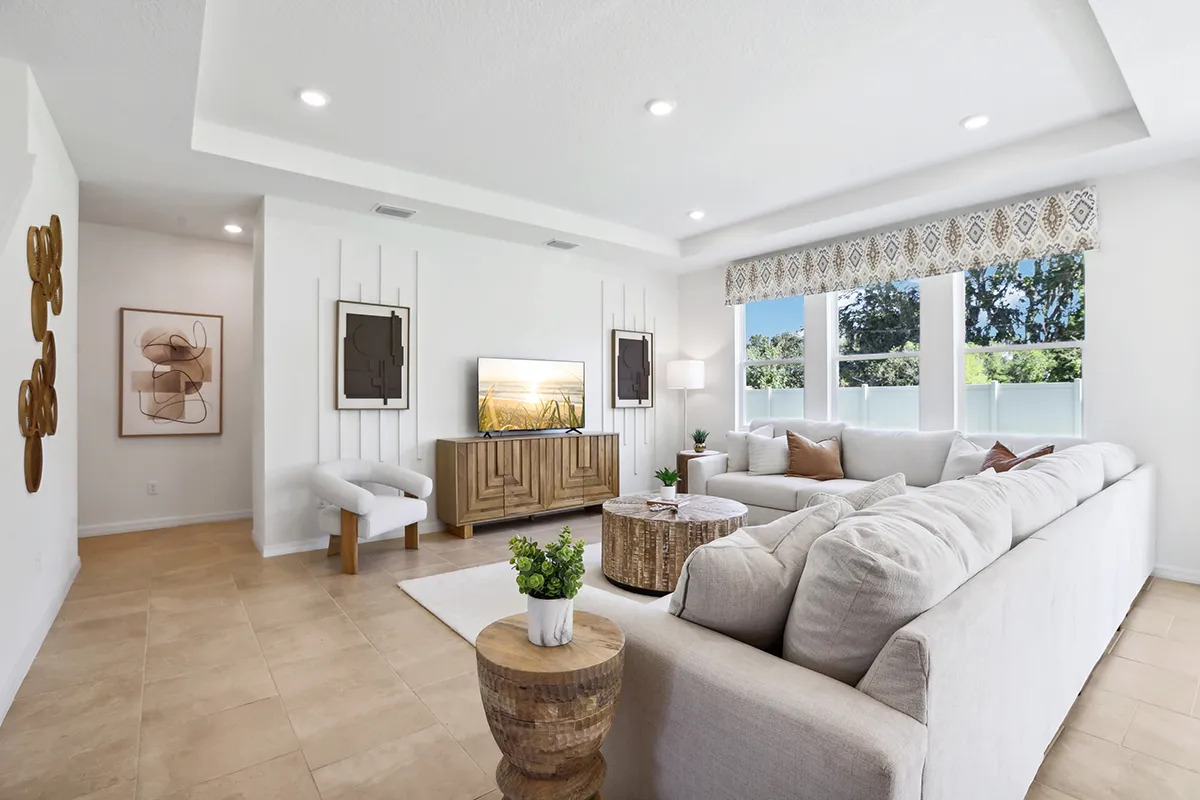 Bright living room with a large white sectional sofa, wooden coffee tables, a TV on a wooden console, modern wall art, large windows with patterned valance, and beige tile flooring.