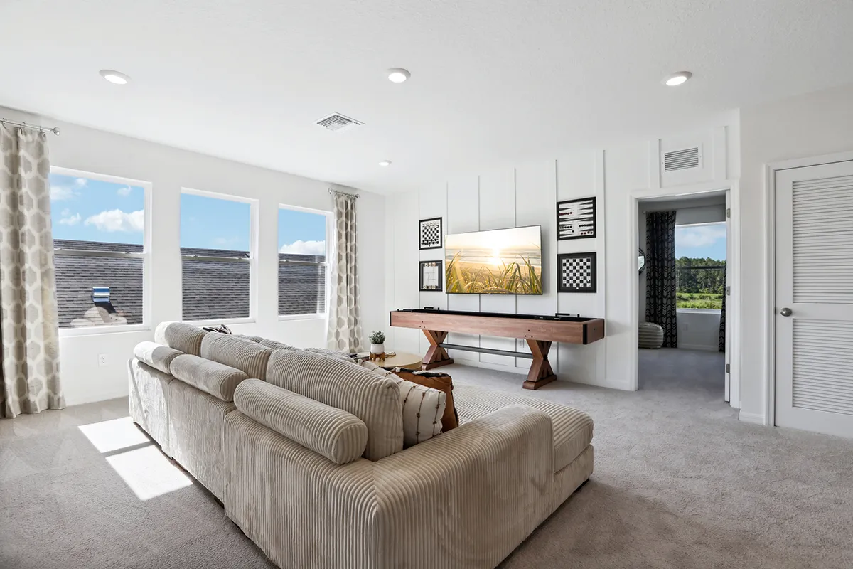 A bright living room with large windows, a beige sectional sofa, light carpet, a wall-mounted TV, patterned wall art, and a console table beneath the TV. A doorway leads to another room with a scenic view.