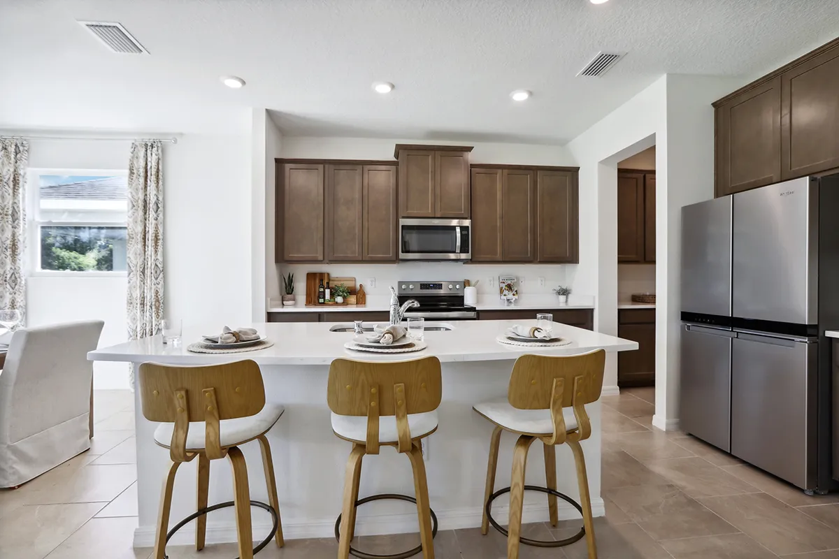 Modern kitchen with wooden cabinets, stainless steel appliances, and a white island with three wooden barstools set with plates and utensils. Natural light enters from a window on the left.