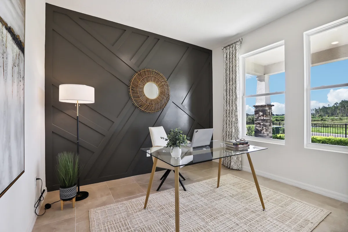 A modern home office with a glass desk, two white chairs, a laptop, floor lamp, and plant. The room has a black accent wall with geometric patterns, round mirror, large windows, and beige tile floor with a rug.