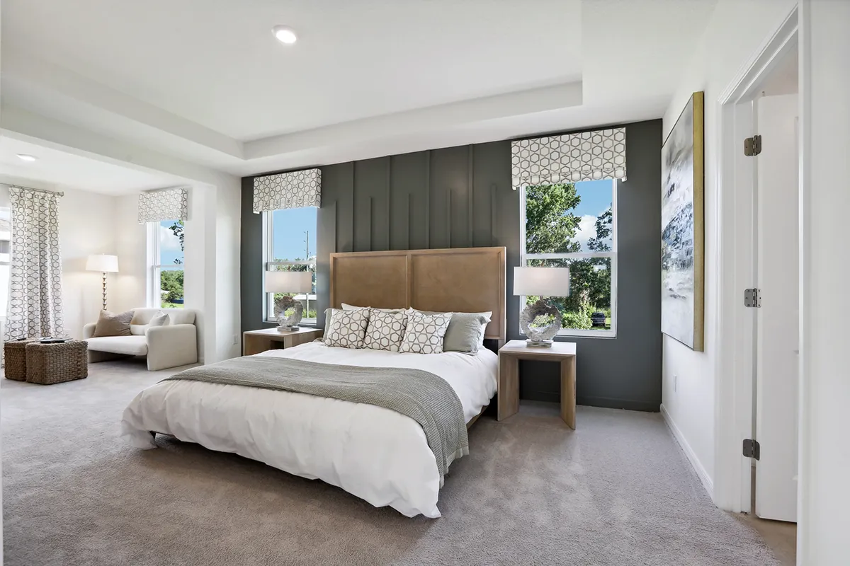 Modern bedroom with a large bed, gray accent wall, two windows, matching nightstands, and contemporary decor. A sitting area with chairs and large windows is visible on the left, with soft natural light filling the room.