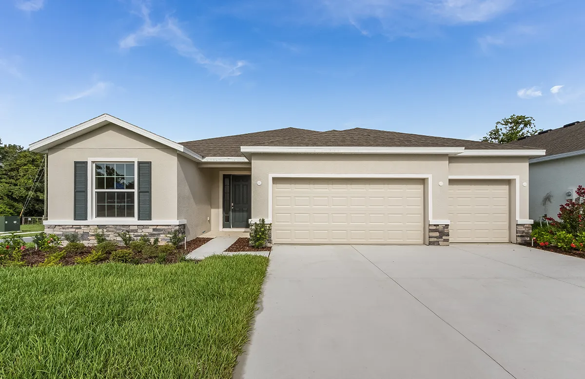 Single-story suburban house with light-colored exterior, dark shutters, and a neatly trimmed lawn. The home features a three-car garage and a wide driveway under a blue sky with scattered clouds.