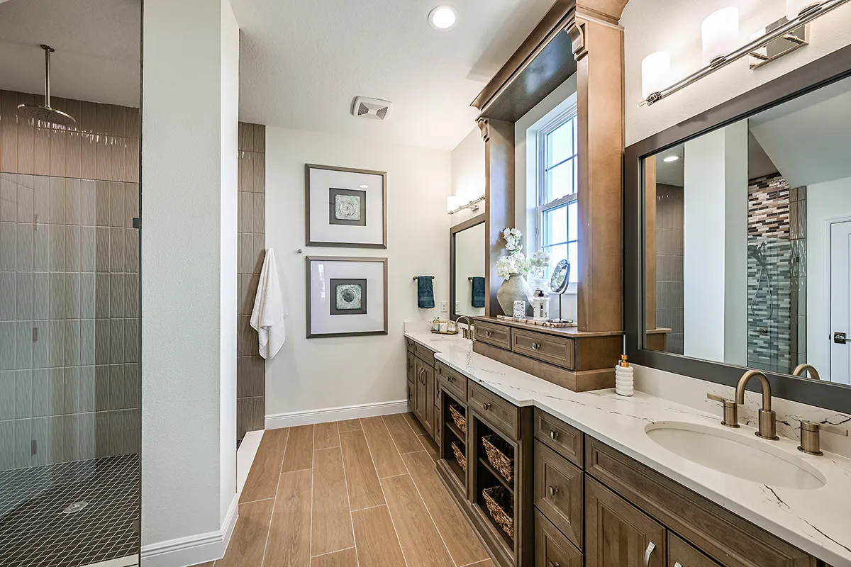 Modern bathroom with a long double vanity, marble countertop, wood cabinets, two sinks, large mirrors, wall lights, and a walk-in shower with tiled walls. Decorative items and framed art hang on the white walls.