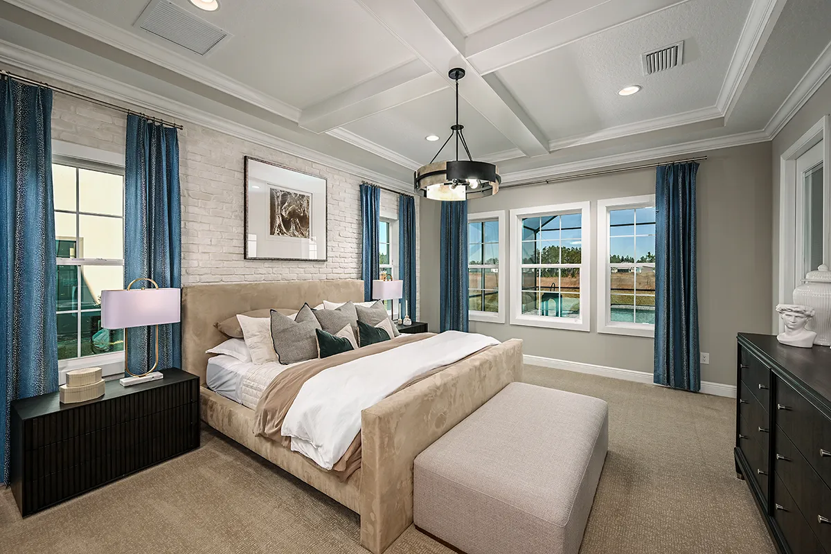 A spacious, modern bedroom with a beige upholstered bed, blue curtains, large windows, a padded bench, two nightstands with lamps, and a dark dresser on the right wall. Light streams in from the windows.