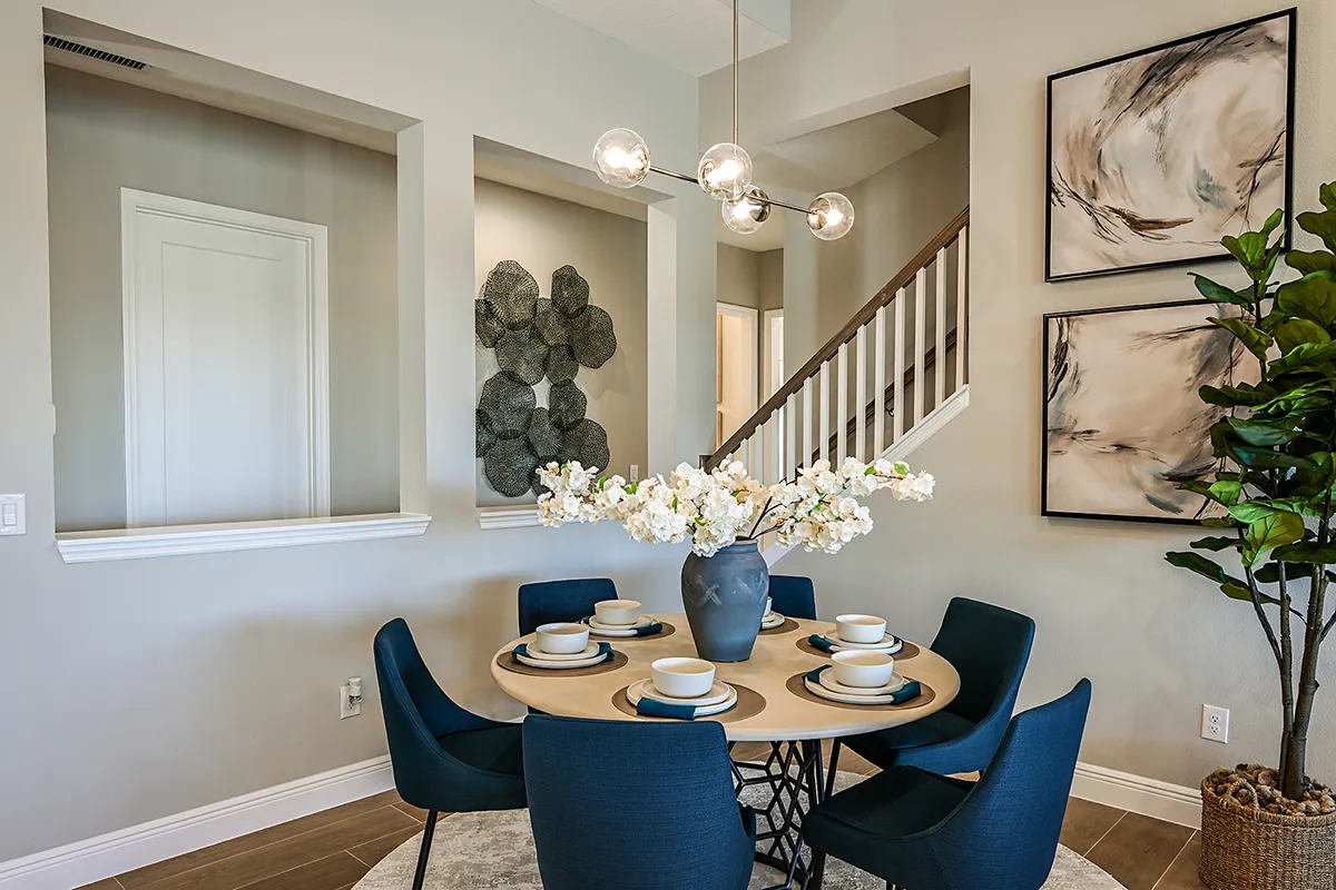 A modern dining area with a round wooden table set for six, navy blue chairs, a vase of white flowers, abstract wall art, a plant, and a staircase in the background. Contemporary decor and neutral tones throughout.