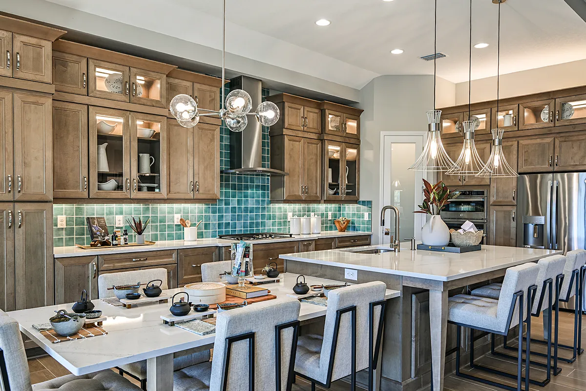 Modern kitchen and dining area with wood cabinets, teal backsplash, stainless steel appliances, two islands with marble countertops, pendant lighting, and beige upholstered chairs. The space is stylish and well-organized.