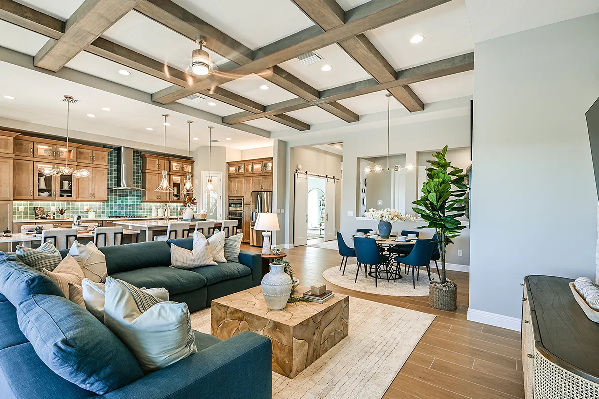 Open-concept living area with a blue sofa, wooden coffee table, and large plant, connected to a modern kitchen with wooden cabinets and a dining area with a round table and blue chairs. Exposed ceiling beams and hardwood floors.