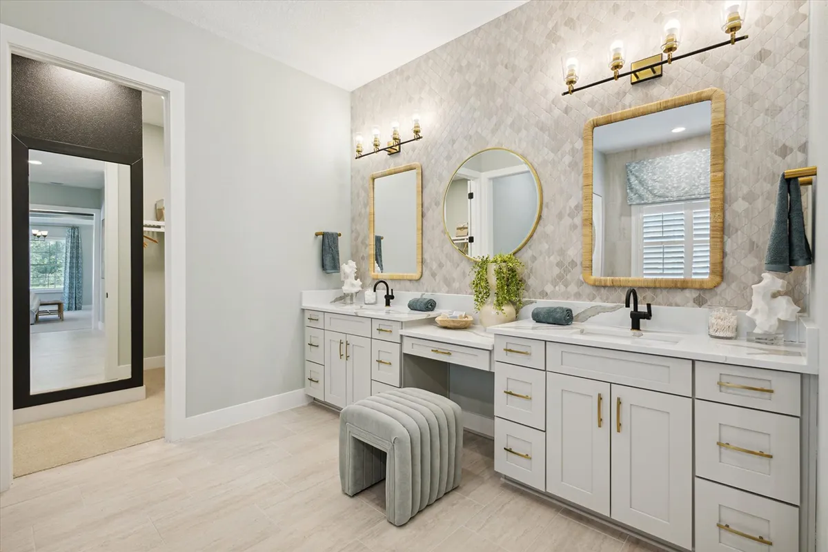 Modern bathroom with dual sinks, white cabinets, gold hardware, two round mirrors, decorative lighting, a cushioned bench, and neutral-toned decor; a doorway opens to a walk-in closet and bedroom beyond.