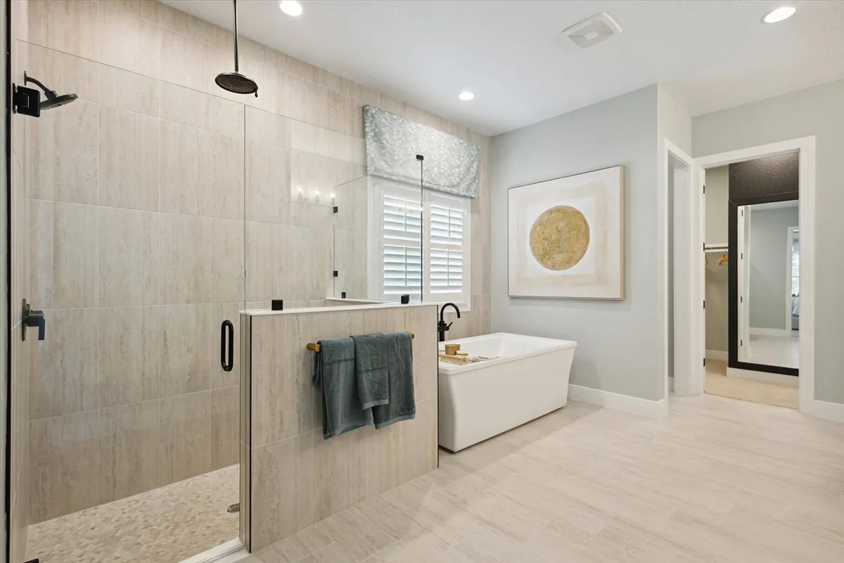 Modern bathroom with a glass-enclosed shower, towel hanging on a half wall, freestanding bathtub, large abstract artwork, light tile floor, and window with white shutters and a gray valance.