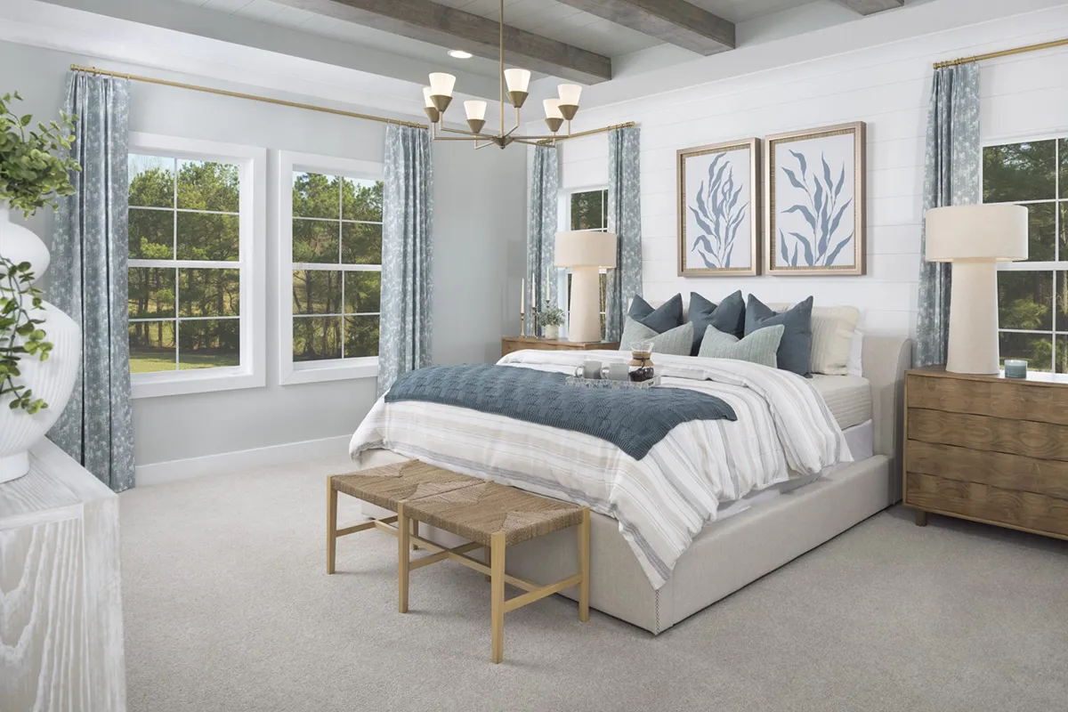 Bright, modern bedroom with a large bed, blue and white bedding, wooden bench at the foot, two nightstands with lamps, large windows with blue curtains, and framed botanical prints above the headboard.