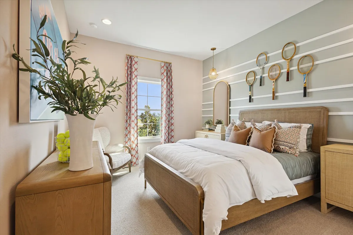 A cozy, modern bedroom with a large bed, neutral linens, wooden furniture, a tall mirror, and decorative tennis rackets on the wall. Sunlight streams in through a window with patterned curtains.