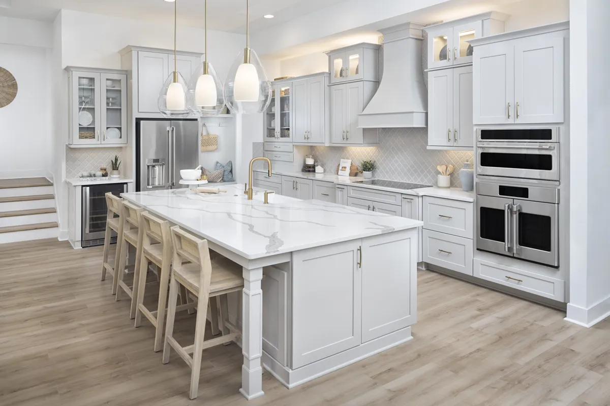 A modern kitchen with white cabinets, a large marble island with four wooden barstools, stainless steel appliances, pendant lights, and light wood flooring. The space is bright and spacious.